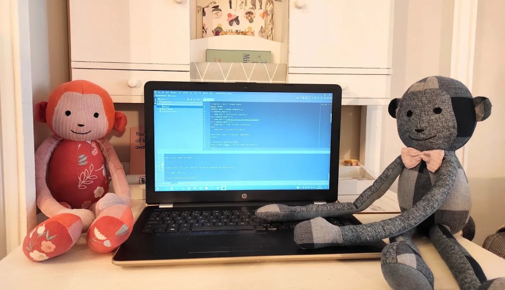 Two handmade toy monkeys sit either side of a laptop. One toy monkey looks like it is coding.