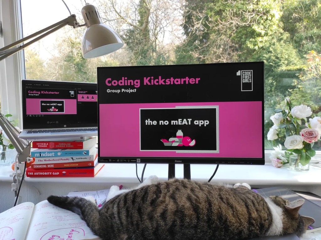 A cat sprawls across my small desk in front of my computer monitor, which displays the presentation for my Code First Girls group project