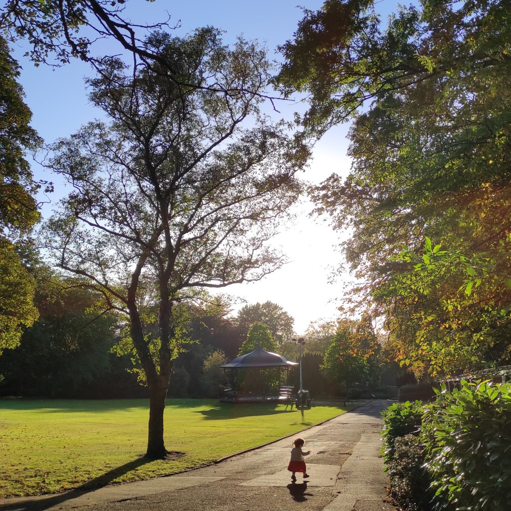My little daughter looks tiny, walking along a path in the park, framed by tall trees, and lit by the setting sun. A sunny post-nursery walk in the park.