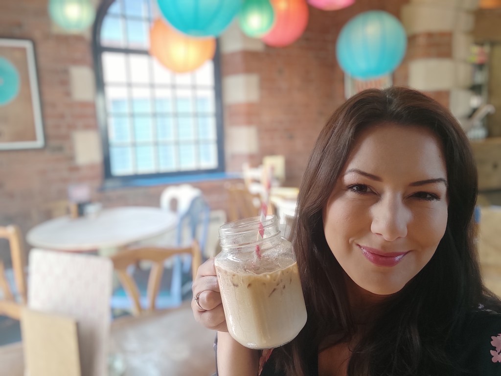 Elena smiles while holding a glass of iced cappucino in a cafe on her first day of work as a UX Designer
